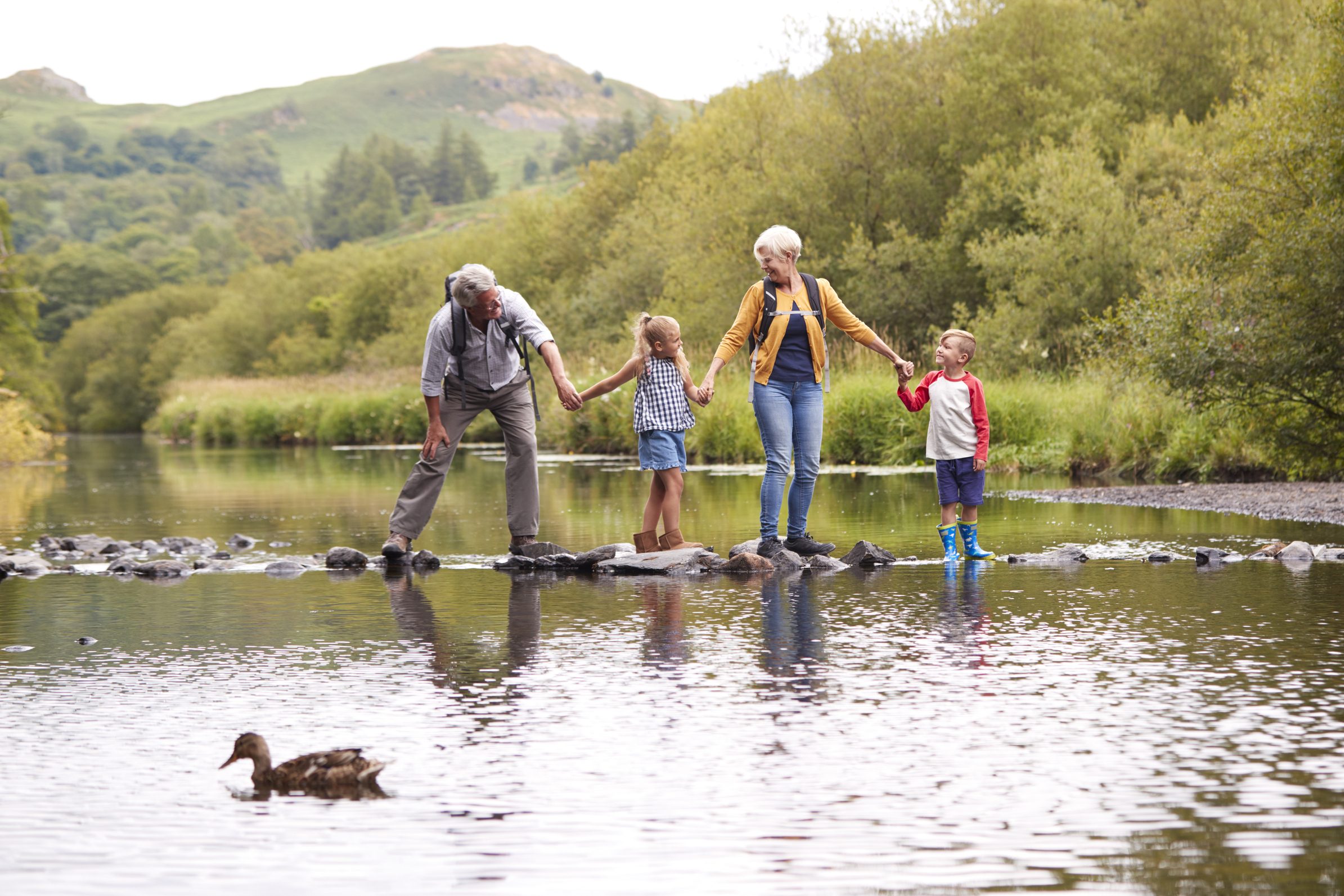 Family crossing river on stepping stones with mountains and duck