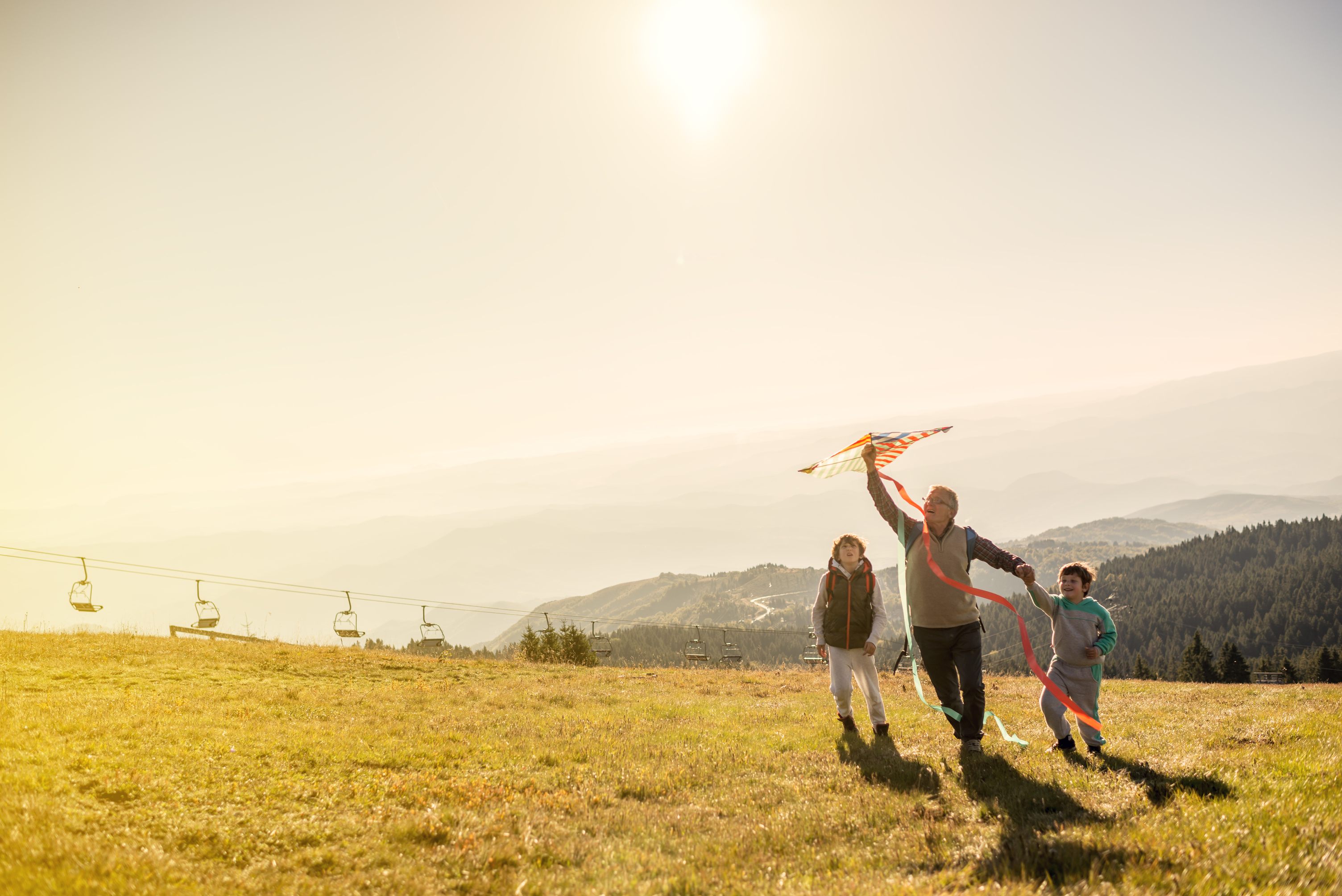 Family flying kite on mountain meadow with ski lifts at sunset