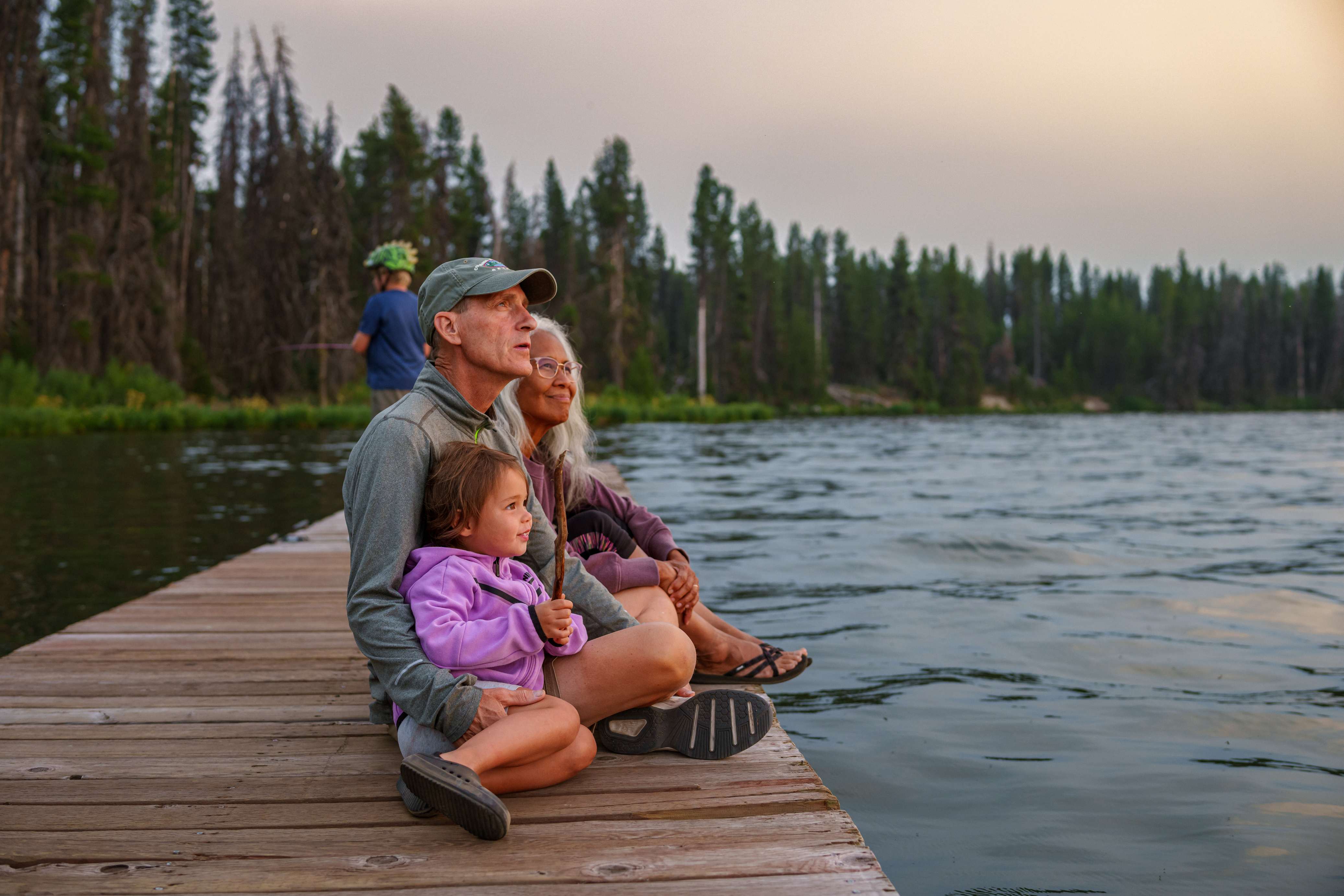 Family sitting on wooden dock by lake surrounded by pine trees