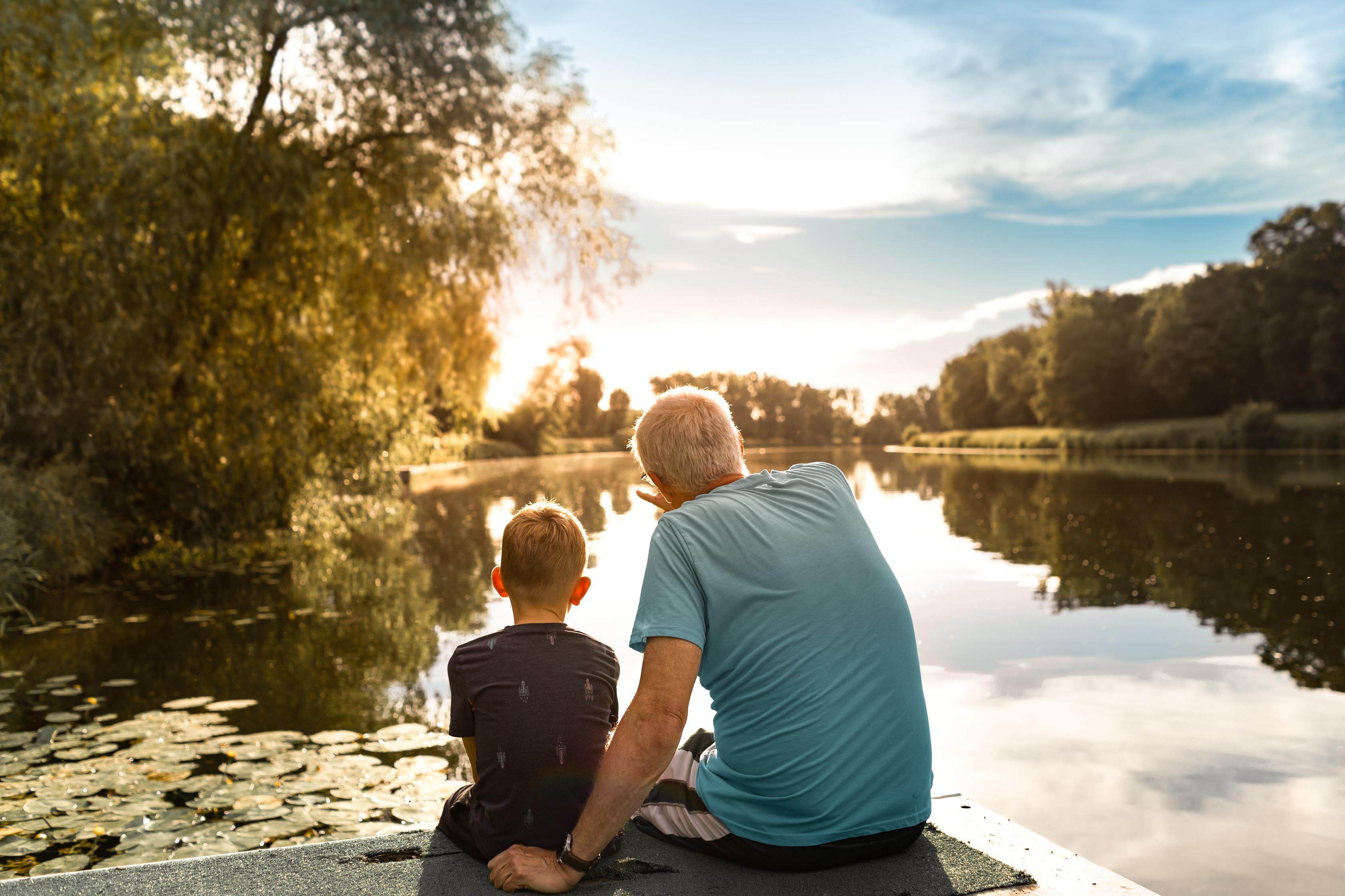 Grandfather and grandson sitting together by a peaceful lake at sunset surrounded by trees