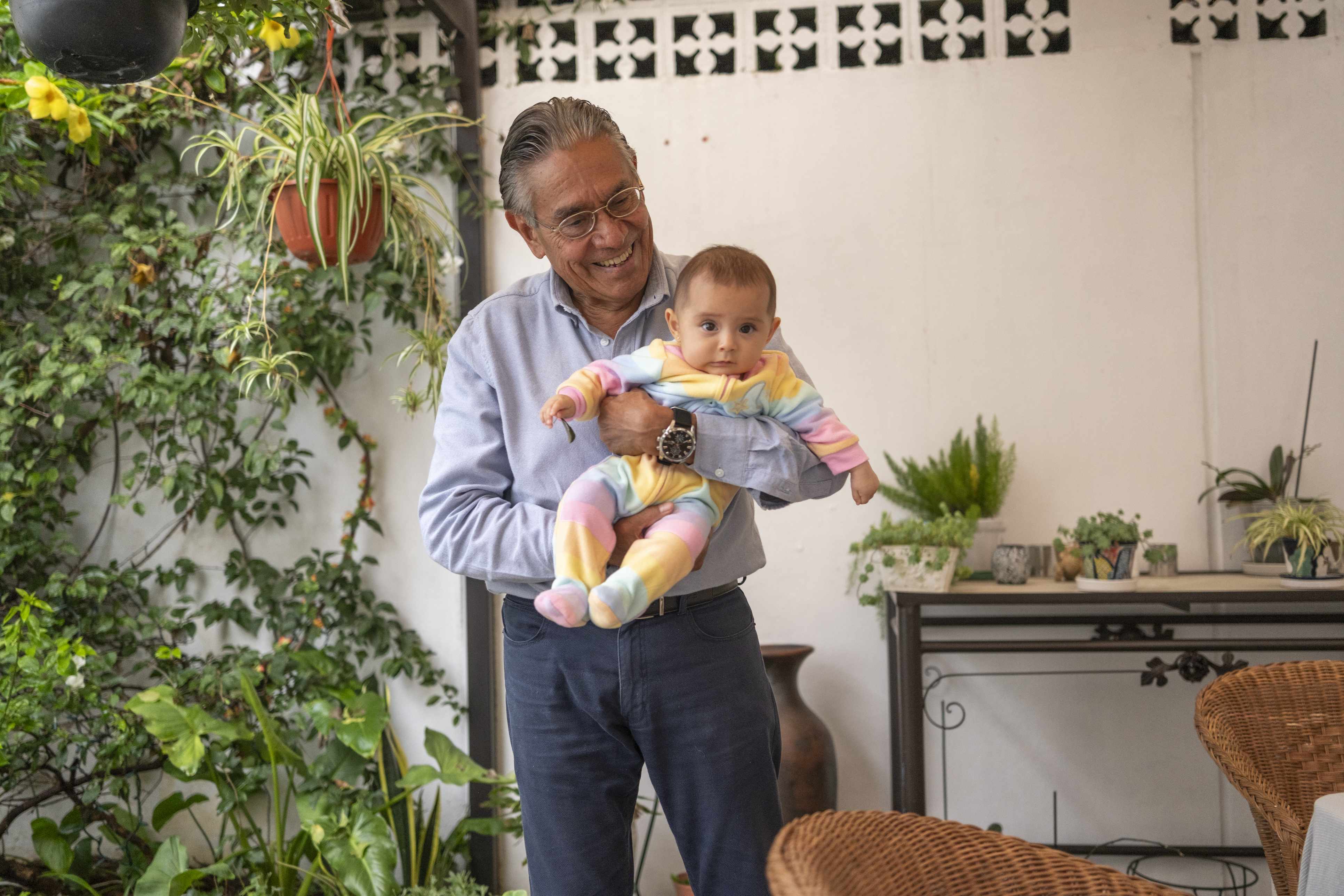 Grandfather holding baby in colorful outfit on outdoor patio with plants