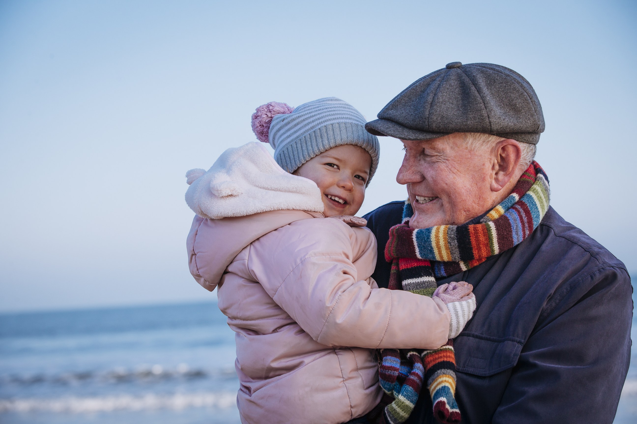 Grandfather holding smiling granddaughter at beach wearing winter clothing