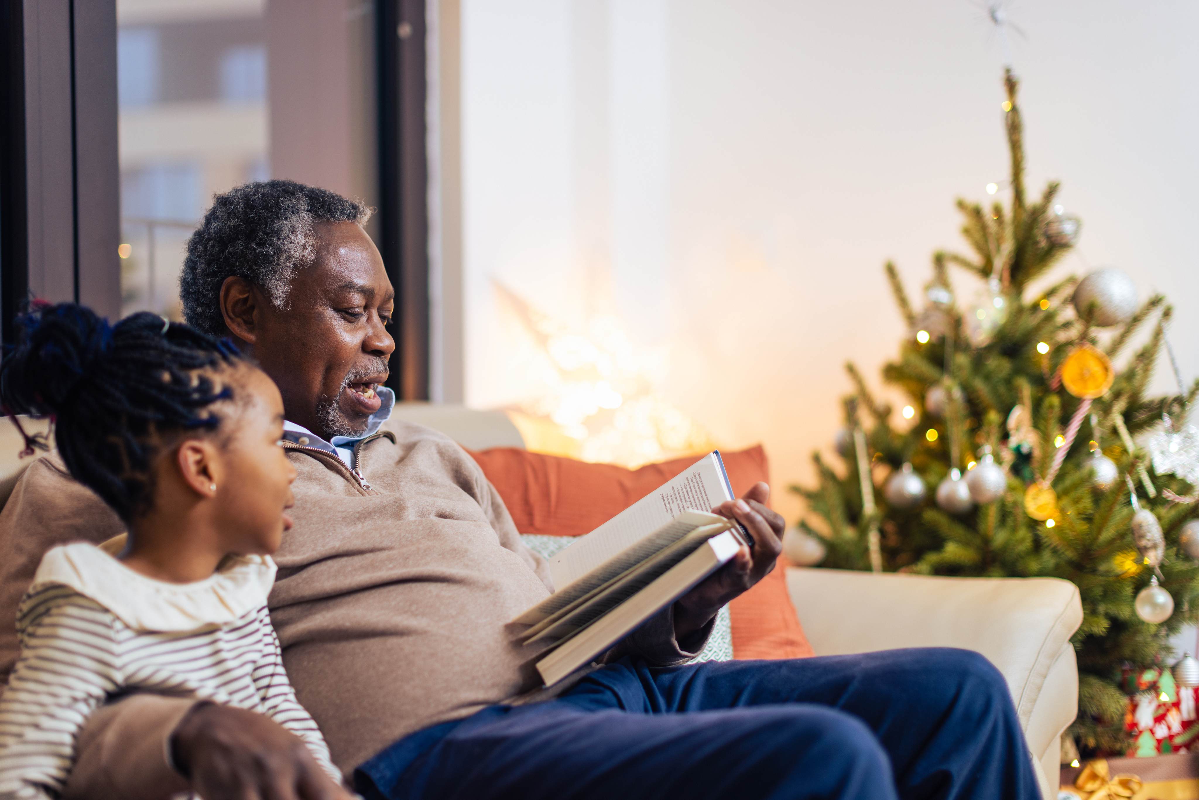 Grandfather reading book to grandchild beside Christmas tree at home