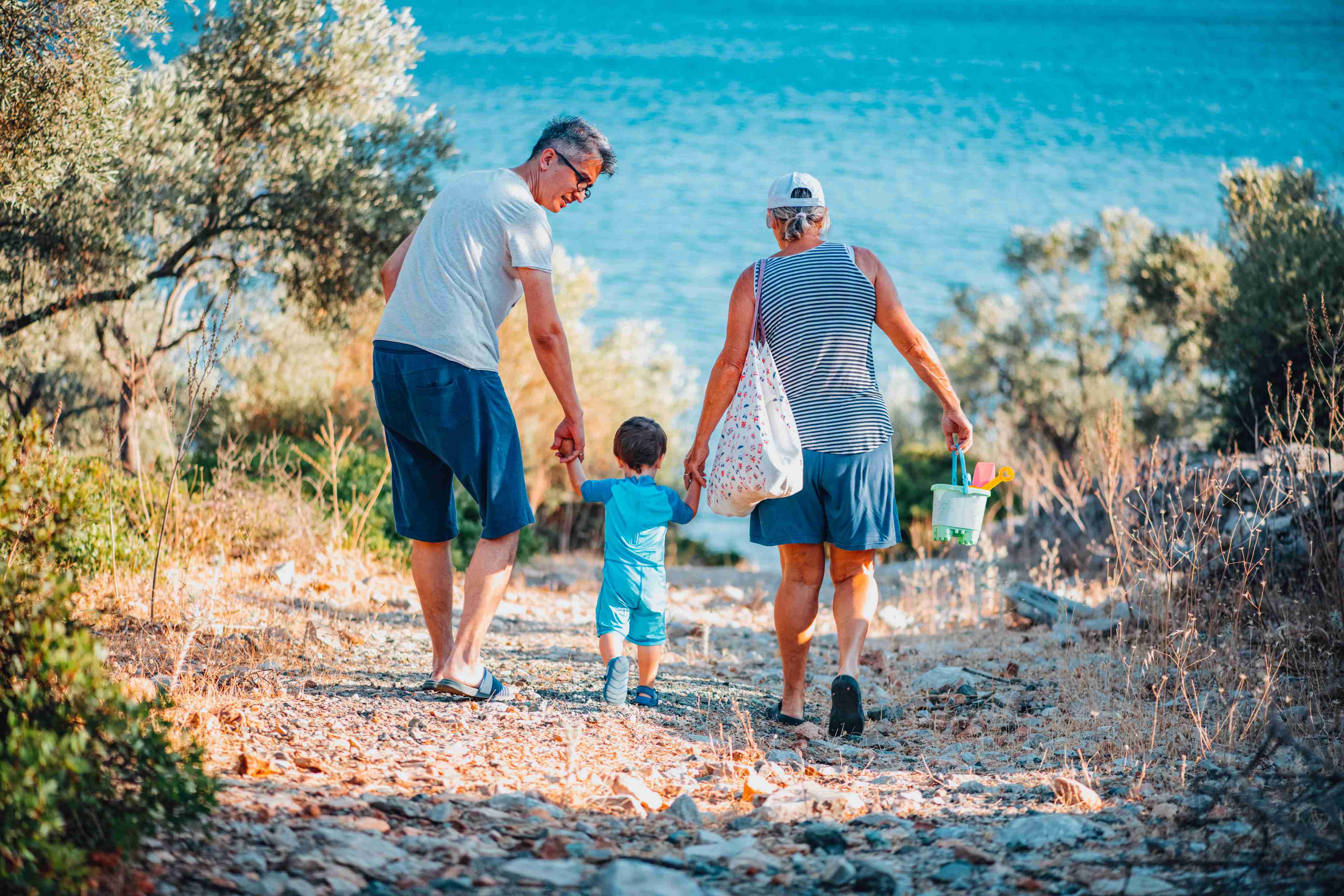 Grandparents walking with young child toward beach carrying beach toys