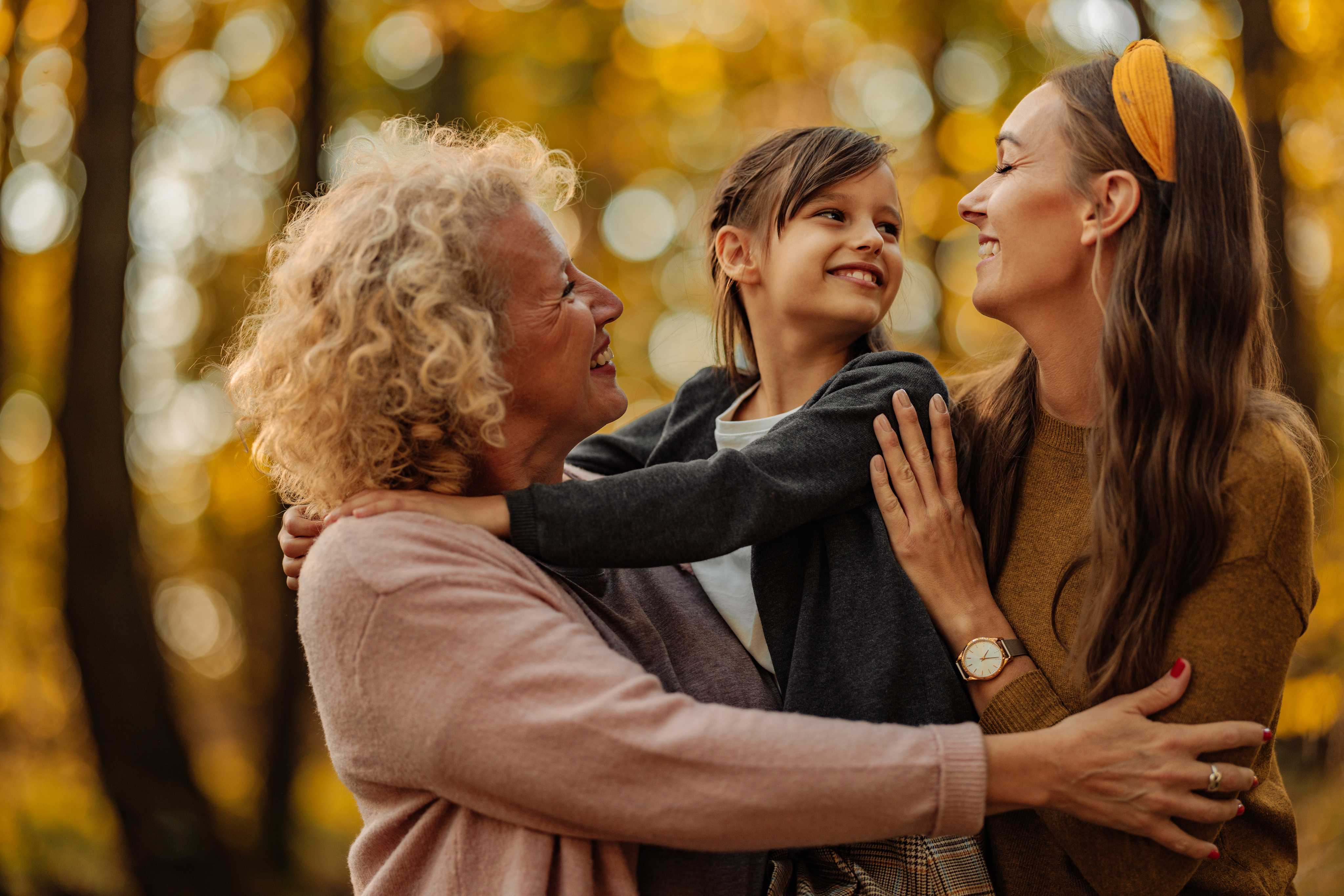 Three generations of women embracing outdoors in autumn with grandmother, mother, and young daughter smiling together against golden fall foliage background
