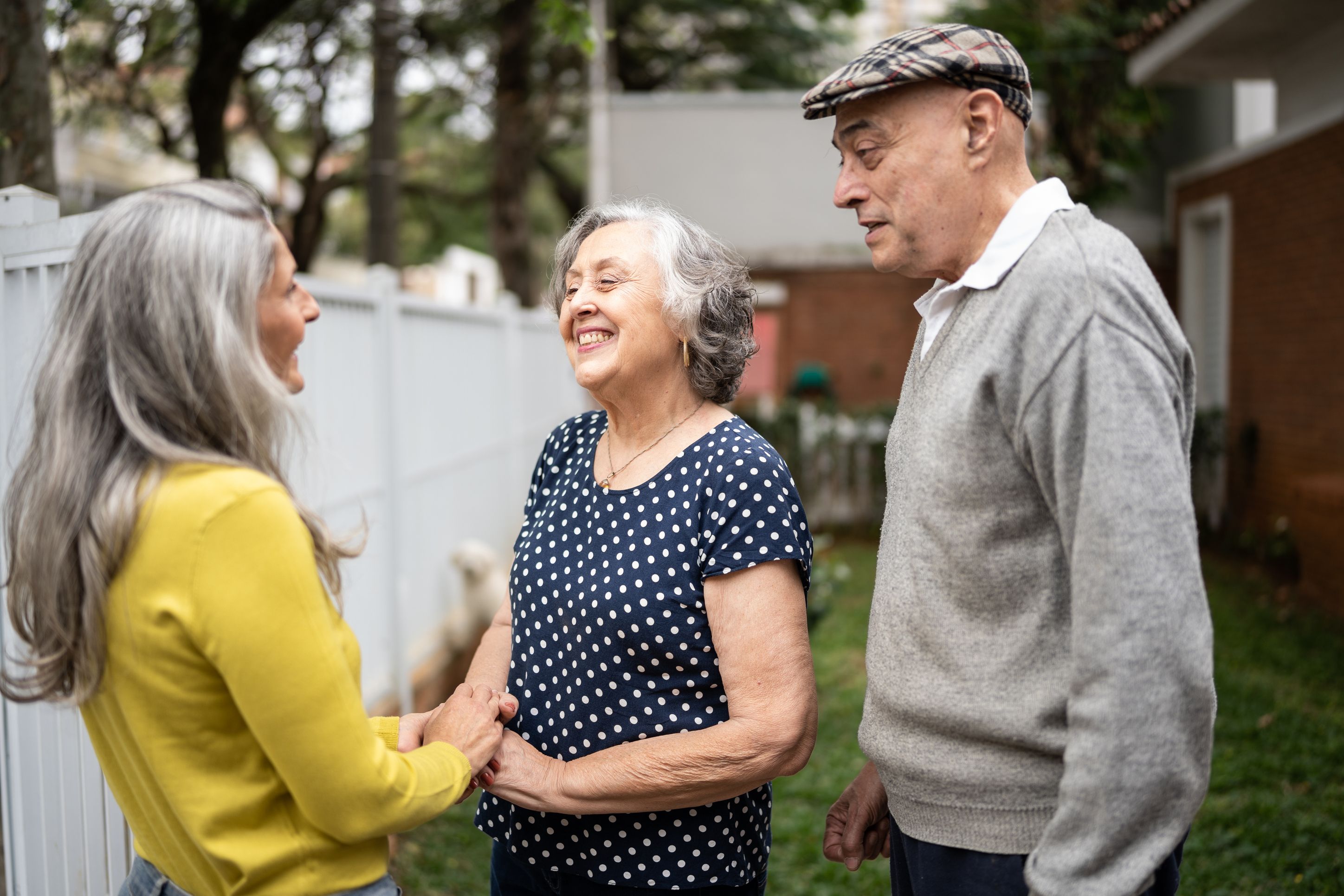 Three older adults having a friendly conversation outdoors, with a woman in yellow talking to a couple, the woman wearing a navy polka dot dress and the man in a gray sweater and plaid cap