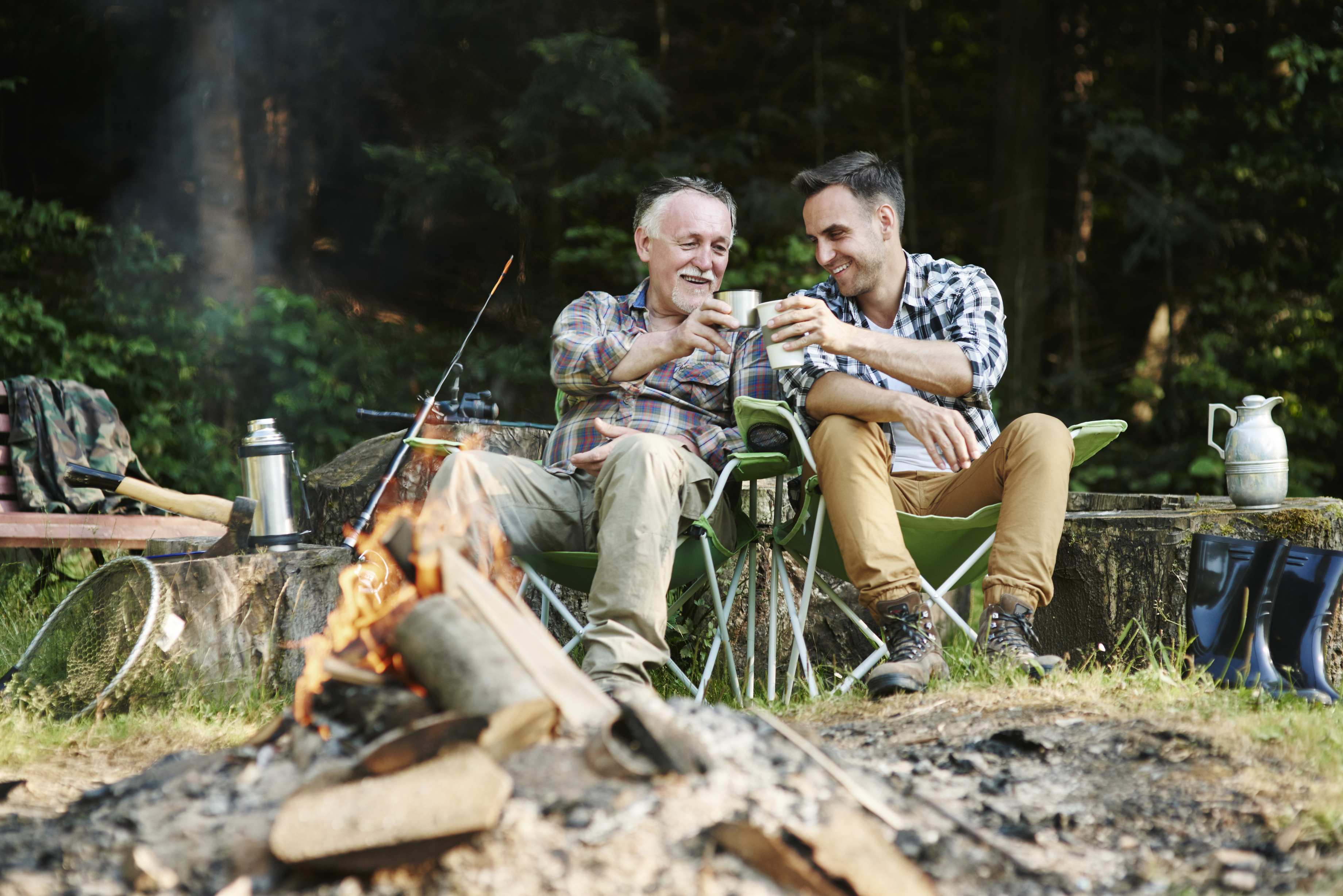 Two men camping outdoors toasting drinks by campfire in forest