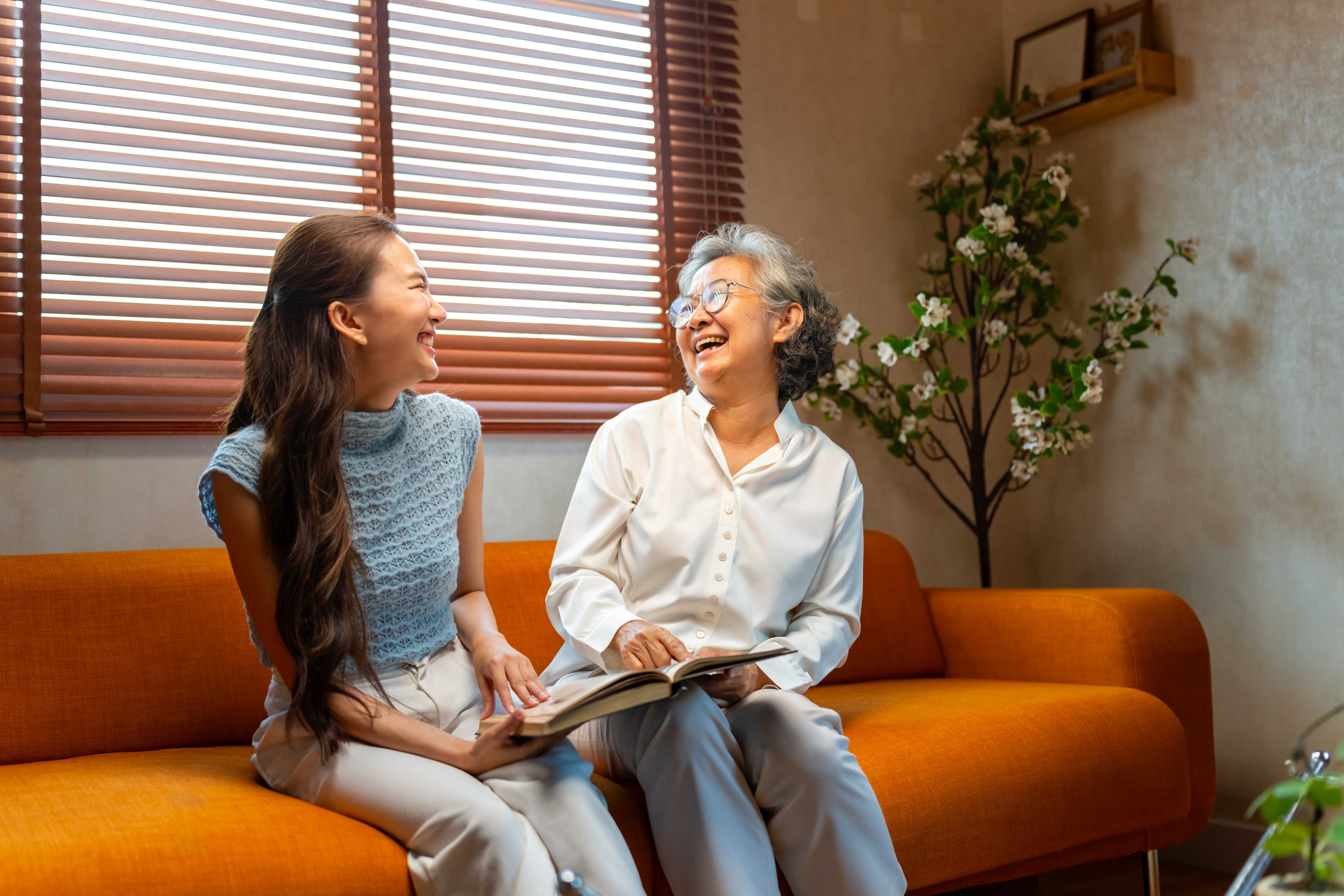 Two women laughing together on orange couch while sharing a book