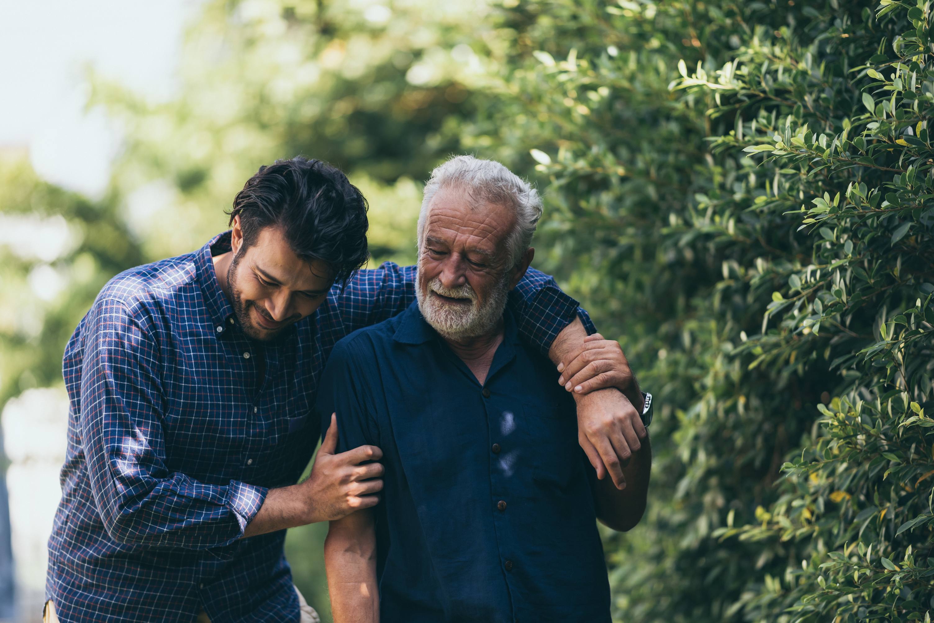Young man in plaid shirt embracing elderly man with gray beard outdoors near green foliage