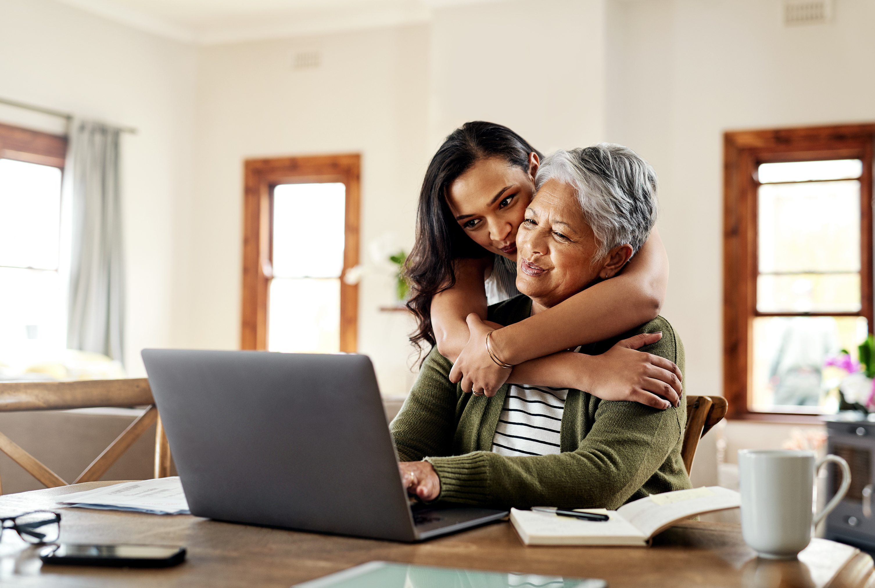 Young woman hugging elderly woman from behind while elderly woman works on laptop at home