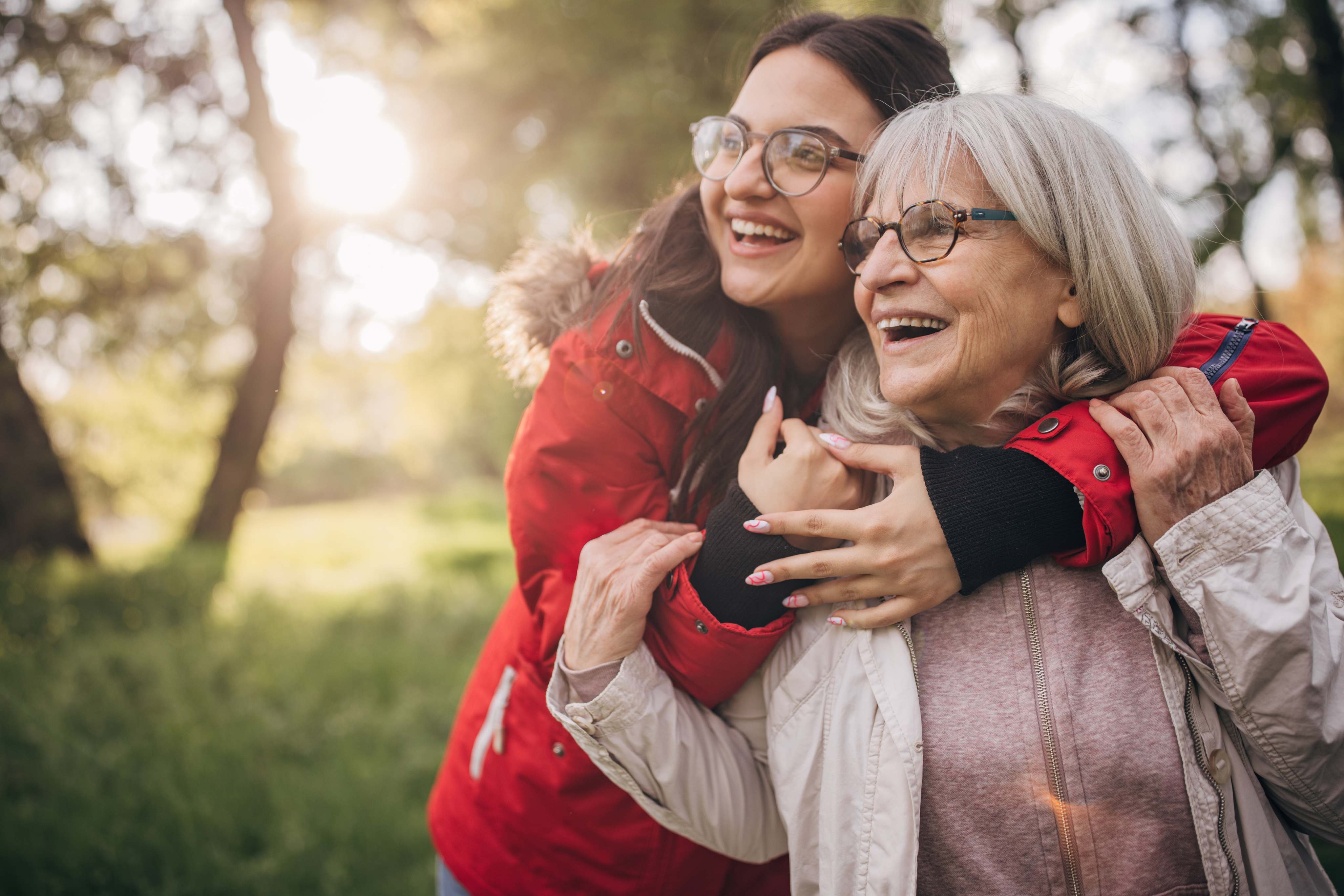 Young woman hugging elderly woman outdoors both smiling and wearing glasses